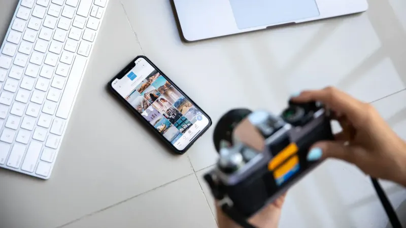 Person using vintage camera with smartphone and laptop in creative setup.