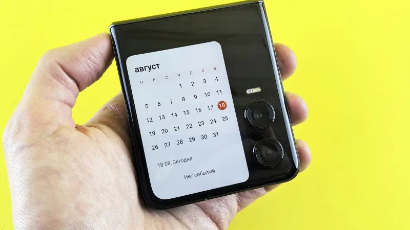 Close-up of a hand holding a flip smartphone with a calendar on screen against a vibrant yellow background.