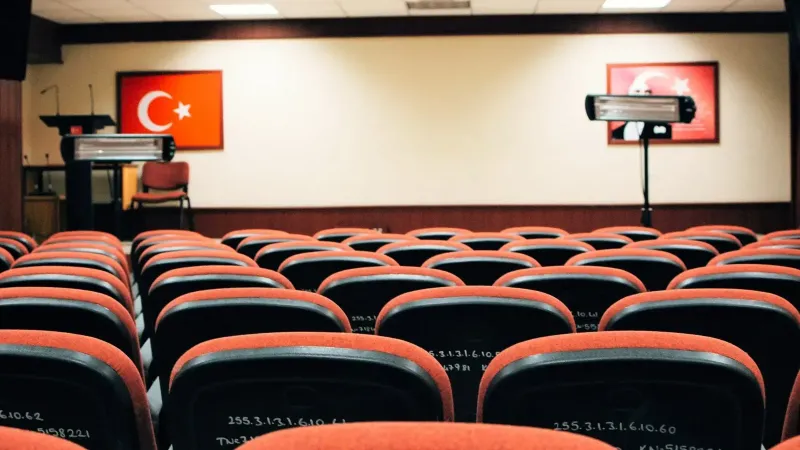 Una sala de conferencias vacía con asientos rojos, un escenario y banderas turcas, lista para una reunión o presentación.