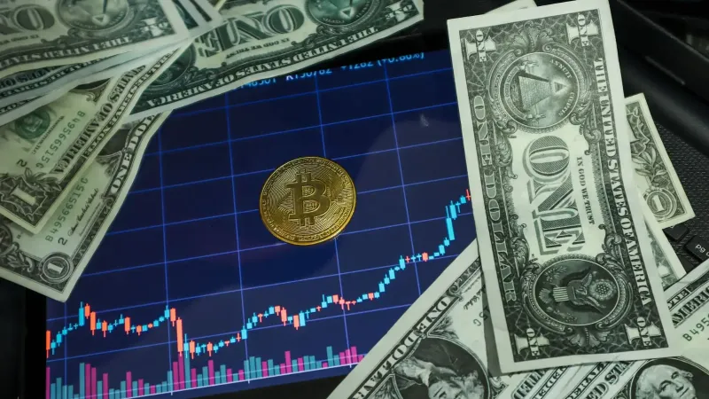 Bitcoin coin on a tablet showing stock chart, surrounded by dollar bills.