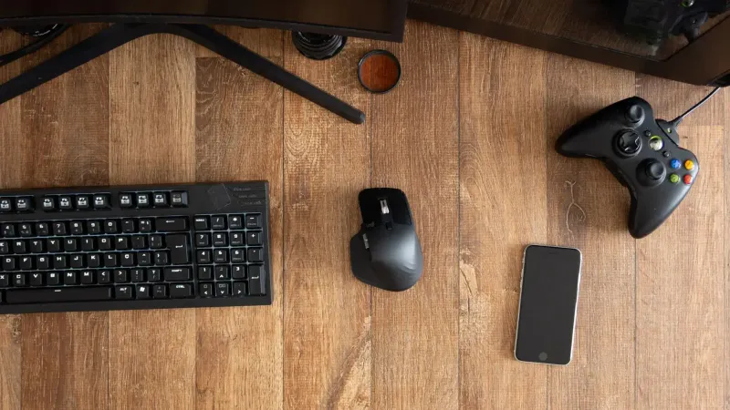 Vista superior del gamepad negro con ratón y teclado en escritorio de madera con monitor en apartamento durante el día