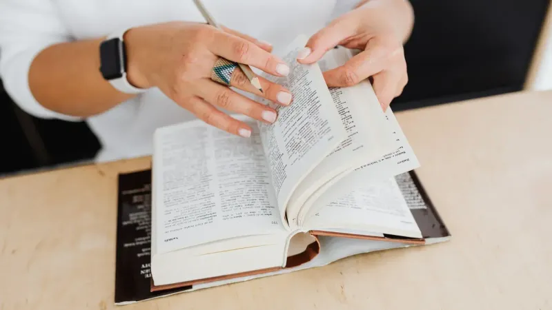 A close-up of a woman turning the pages of a dictionary on a wooden desk.
