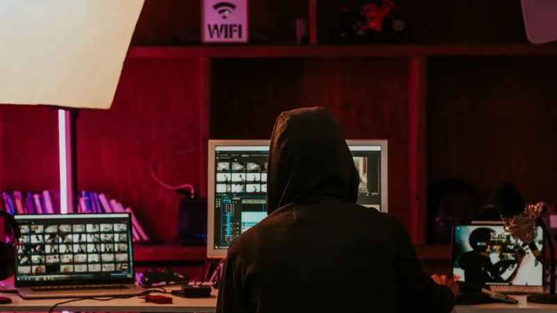 Person in a hoodie editing videos on multiple screens in a dimly lit space with neon lighting.