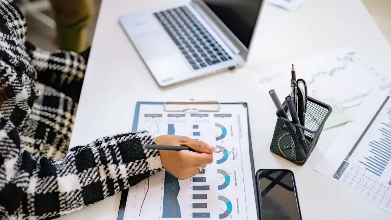 An office setting with a person analyzing charts and graphs at a desk with a laptop and smartphone.