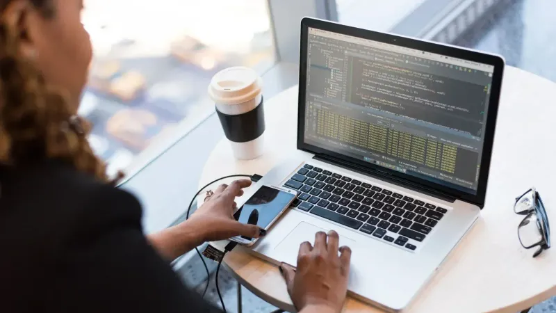 Mujer negra programación en una computadora portátil con café, teléfono inteligente y gafas en un escritorio en una oficina.