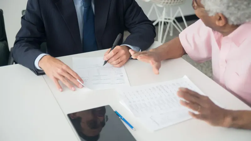 A business meeting between an elderly client and a consultant discussing documents at an office table.