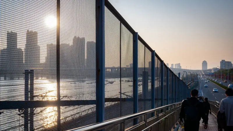 Silhouetted cityscape during sunset with people walking along a fenced bridge.