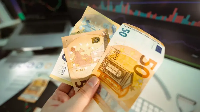 Hand holding Euro banknotes in front of a trading screen showing financial graphs.
