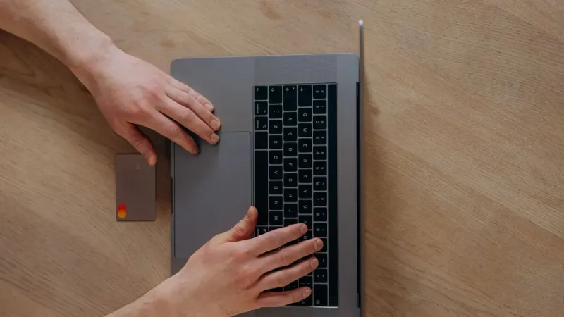 A person using a laptop and credit card for online shopping, viewed from above on a wooden table.
