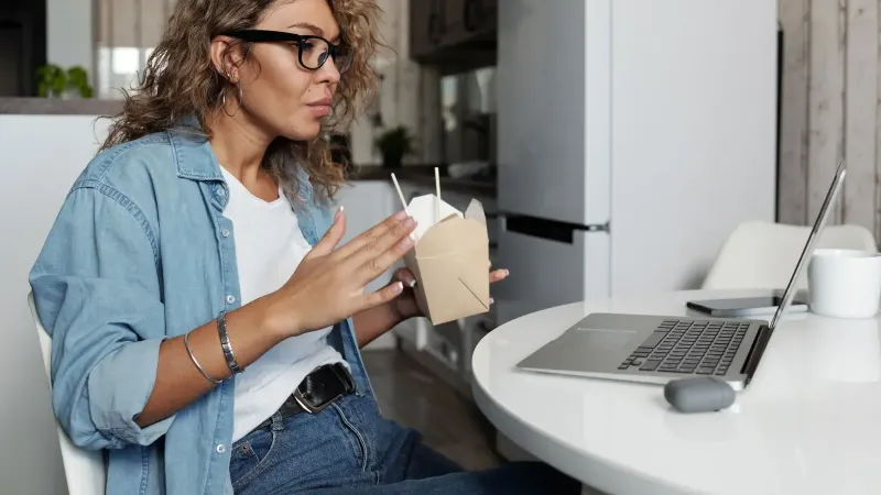 Una mujer enfocada en un ambiente acogedor en el hogar, trabajando en una computadora portátil mientras disfruta de la comida para llevar.