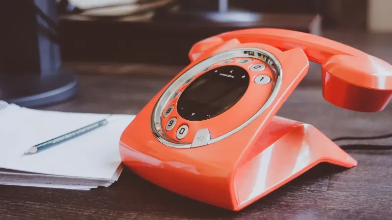 Un teléfono giratorio naranja de estilo retro se encuentra en un escritorio de madera junto con un lápiz y un bloc de notas.