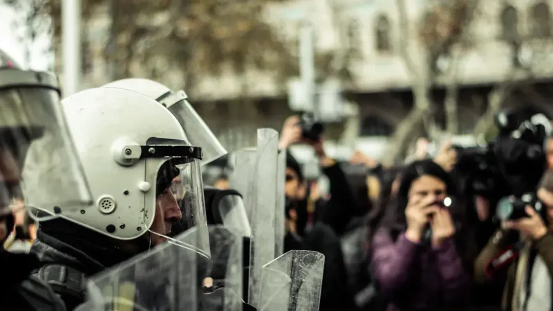 Police officers in helmets and shields confronted by crowd of photographers.