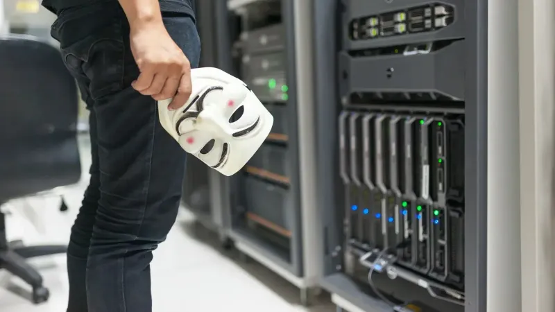 Persona que sostiene una máscara anónima cerca de servidores, insinuando la seguridad cibernética y hackeando temas.
