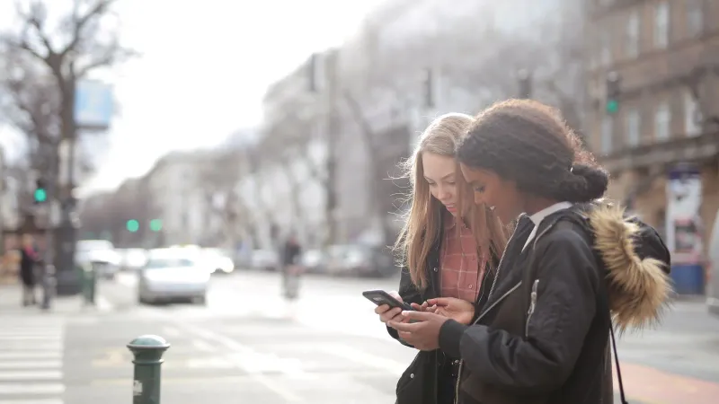 Two young women looking at a smartphone on a bustling urban street during the day.