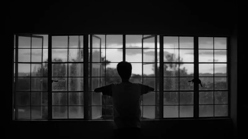 A moody silhouette of a man staring out an open window, evoking solitude.