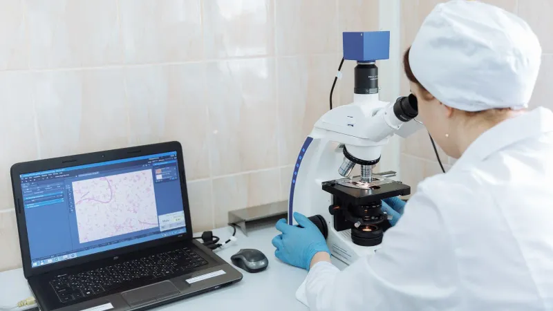 Scientist in lab coat using microscope and laptop in a laboratory setting.