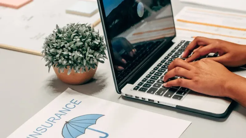 Hands typing on a laptop at a desk with an insurance paper and plant, suggesting a work environment.