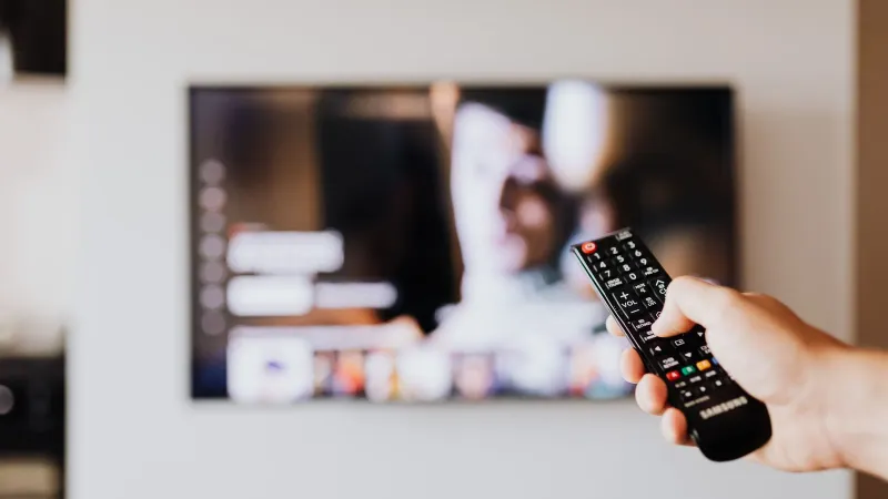 Person using a TV remote control with a blurred television screen in the background.