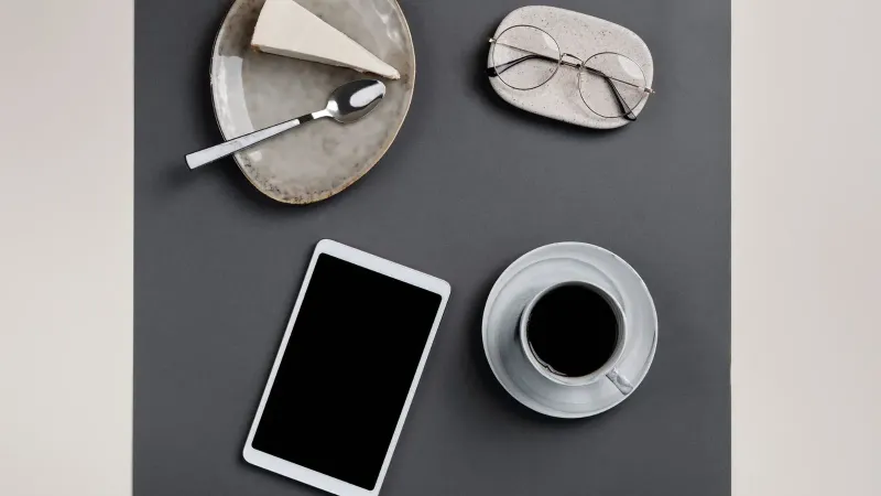 Stylish flat lay featuring coffee, cheesecake, eyeglasses, and a tablet on a gray table, perfect for lifestyle themes.