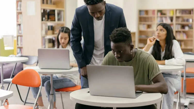 Students learning in a classroom setting with a teacher assisting and laptops on desks, creating an interactive education environment.