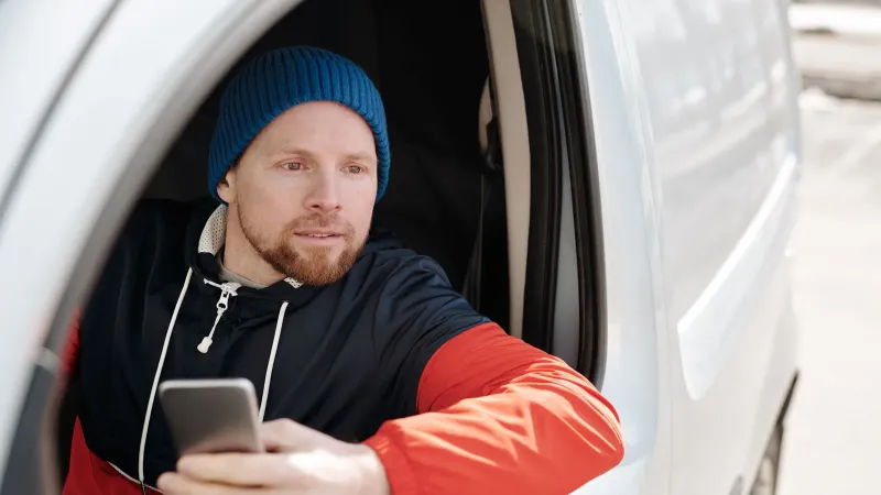 Stylish man in a car looking out the window holding a smartphone, wearing a beanie.
