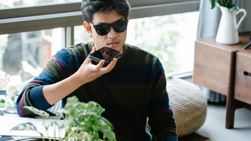 A man wearing sunglasses uses a smartphone's voice assistant in a cozy indoor setting.