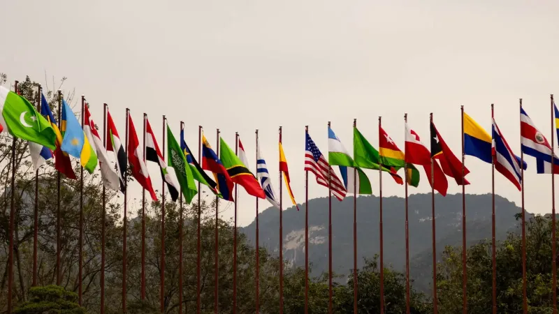 A display of various national flags on flagpoles set against a mountain backdrop, symbolizing global unity.