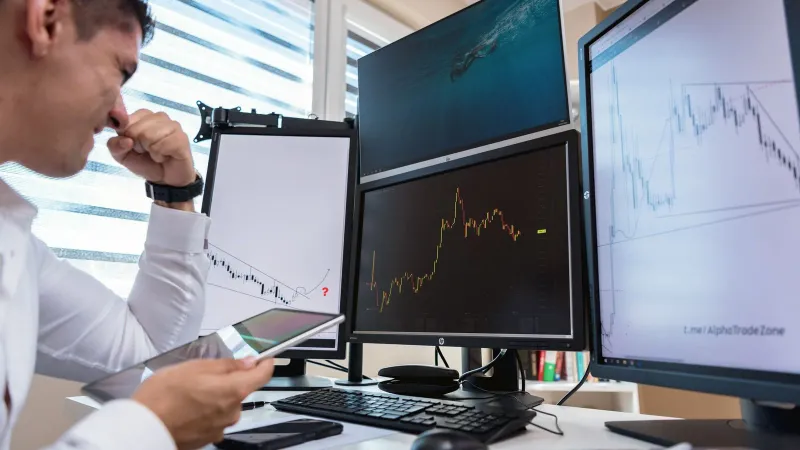 Trader in white shirt analyzing stock charts on multiple monitors during daytime in an office setting.