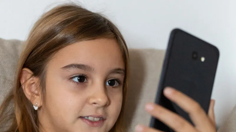 A young girl with long brown hair smiles while using a smartphone indoors.