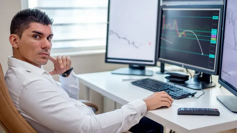Professional stock market analyst monitoring data on multiple screens at a workstation.