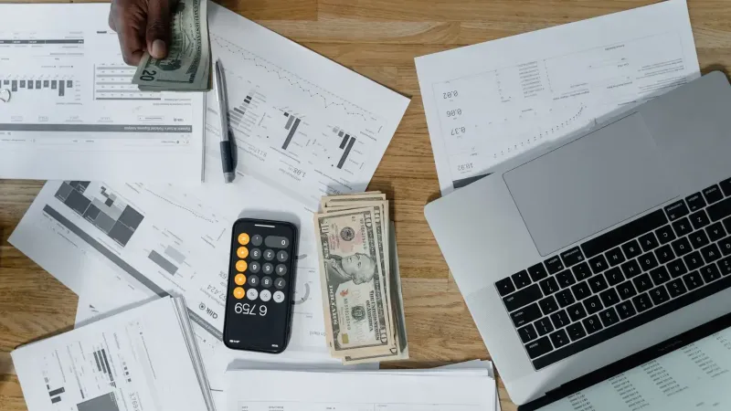 Overhead view of a busy workspace with cash, financial documents, and a laptop.