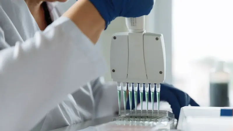 A scientist wearing a lab coat uses a multichannel pipette for liquid handling in a lab setting.