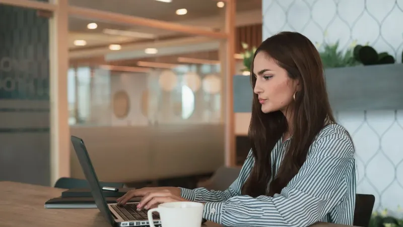 A professional woman working on her laptop at a modern office with a coffee cup nearby.