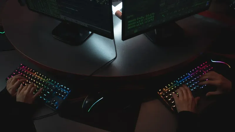 Two people typing on RGB keyboards with code on screens, indicating a cybersecurity environment.