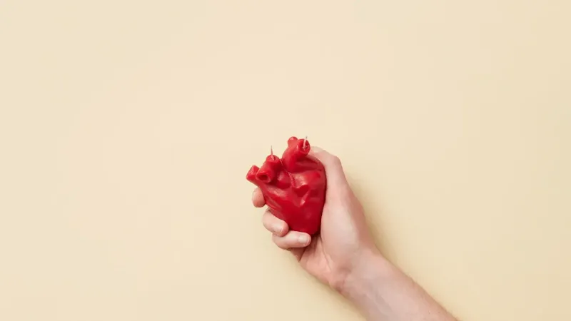 Un modelo de corazón anatómico rojo realista sostenido firmemente en una mano sobre un fondo beige.