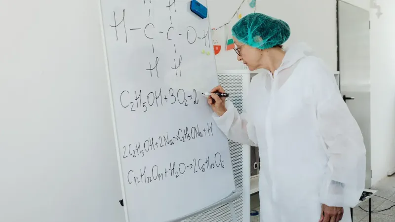 Anciana en bata de laboratorio escribiendo ecuaciones químicas en una pizarra en el interior.