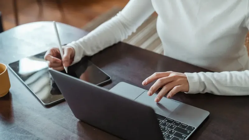 From above of crop anonymous businesswoman in white sweater working at table using tablet with stylus and typing on laptop