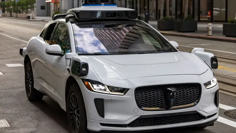 A white autonomous vehicle navigating a city street, reflecting urban architecture in daylight.