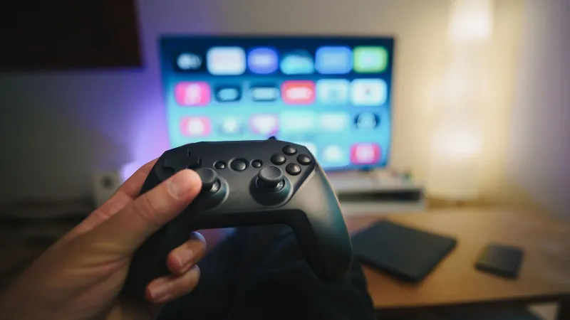 Close-up of a person holding a gaming controller in a cozy living room setup with TV.