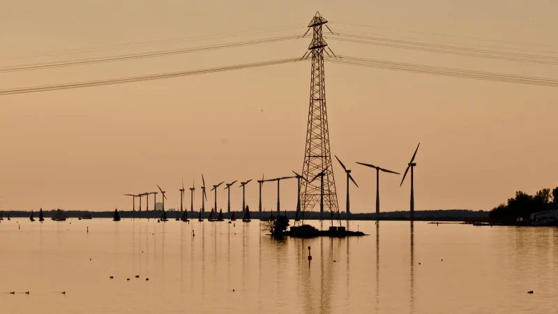 Silhouette of wind turbines and power lines over water during sunset, reflecting sustainable energy.