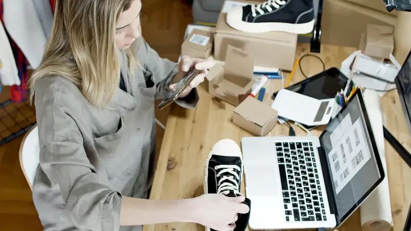 Mujer fotografiando zapatos para la venta en línea en el espacio de trabajo del hogar con laptop y paquetes.