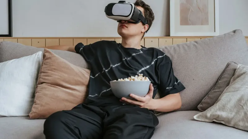 Man sitting on a sofa wearing a VR headset and holding a bowl of popcorn, enjoying technology.