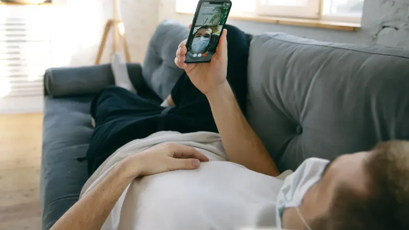A man lying on a sofa using a smartphone for a video call while wearing a face mask indoors.