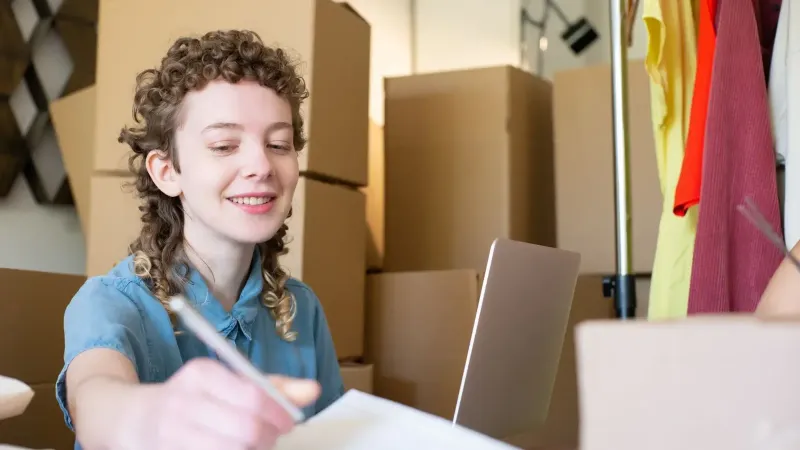 Mujer joven sonriente que gestiona una pequeña empresa con laptop y paquetes a su alrededor.