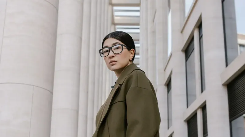 Portrait of a stylish woman in eyeglasses, olive coat, against modern architectural backdrop.