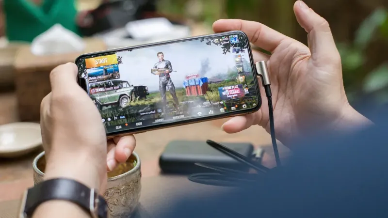Person playing PUBG on smartphone outdoors with charging cable attached.