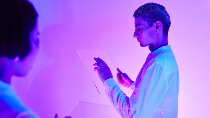 Two individuals engaging with futuristic transparent touch screens in a vibrant purple-lit studio.
