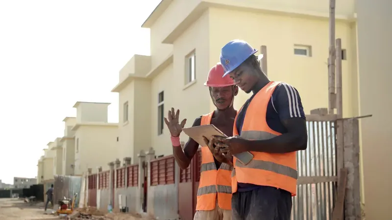 Dos trabajadores de la construcción en cascos y chalecos revisan planos en un sitio.