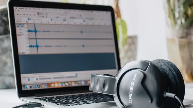 Primer plano de una computadora portátil con software de edición de audio y auriculares en un entorno de oficina en casa.
