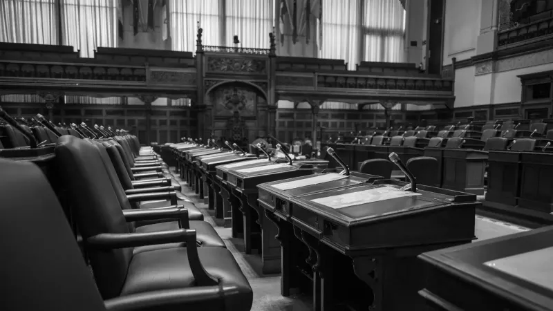 Foto en blanco y negro de una cámara de asamblea legislativa tradicional, con asientos y escritorios vacíos.